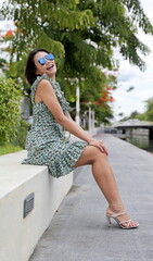 Joyful Asian woman sits confidently, smiling, wearing a chic floral-patterned dress with ruffled details and beige high-heeled sandals