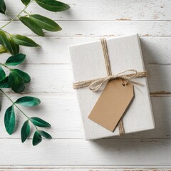 A simple white gift box with a kraft tag, tied with twine, sits on a white wooden surface with greenery