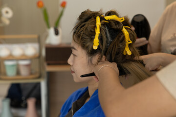 Asian woman getting her hair styled at salon, hairdresser using comb and hairdryer, hair clips...
