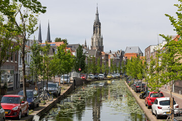 Naklejka premium A picturesque Delft canal view, featuring the majestic Nieuwe Kerk spire, charming canal-side houses, and parked cars under a sunny sky. Delft, Netherlands, 31 May 2025.