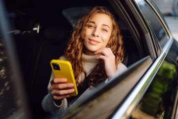 Young woman with phone sitting on back seat of car looking out window. Happy woman in taxi texting...