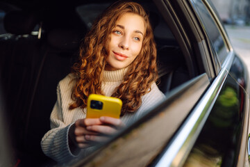 Young woman with phone sitting on back seat of car looking out window. Happy woman in taxi texting...
