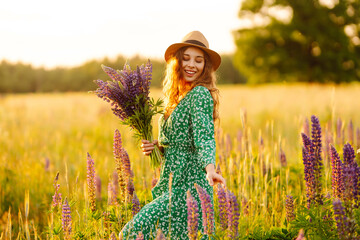 Cute woman in green dress holding beautiful bouquet of lupines on sunset field. Beautiful woman...