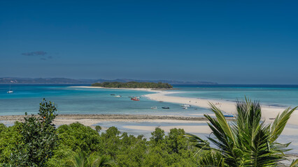 Beautiful tropical landscape. A postcard view. A sand spit  winds to a small green island. Boats in the aquamarine ocean. Silhouettes of people on the beach. Green vegetation, palm trees. Madagascar.