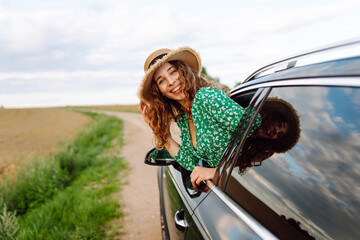 Traveler in hat and bright dress leaning out of car window. Beautiful woman in hat enjoying car...