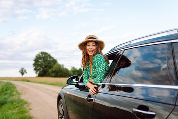 Traveler in hat and bright dress leaning out of car window. Beautiful woman in hat enjoying car...