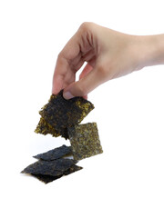 A hand picking up a square piece of dried seaweed snack with a stack of seaweed sheets on a white background, isolated for commercial food presentation or packaging use.