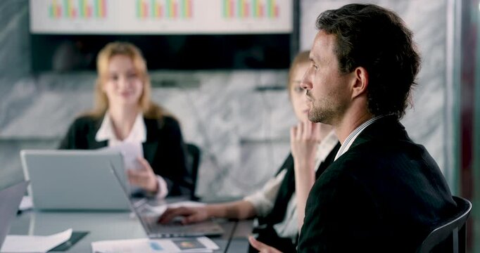 Caucasian male adult employee listening carefully during office meeting with colleagues exchanging ideas using laptops discussing teamwork strategy and presenting data on large wall chart behind team