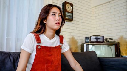 Beautiful young asian woman is sitting at sofa in home holding digital tablet. She is tired and...