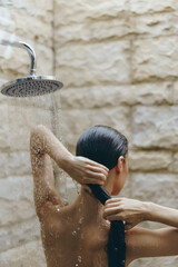 woman showering in outdoor bathroom with stone wall, wet hair, natural light, relaxing self-care moment, wellness and hygiene routine
