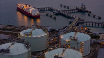 Aerial view of an LNG tanker docked at an offshore terminal with multiple pipelines, storage tanks, and an industrial complex. Captures global energy logistics