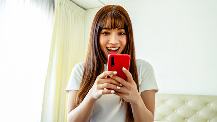 Happy asian young woman sit on bed at home typing on mobile phone receive great news celebrating...