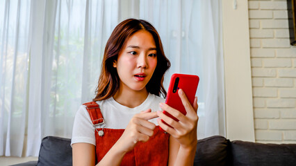Happy asian young woman sit on sofa at home typing on mobile phone receive great news celebrating...
