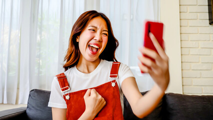 Happy asian young woman sit on sofa at home typing on mobile phone receive great news celebrating...