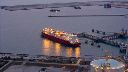 Aerial view of an LNG tanker docked at an offshore terminal with multiple pipelines, storage tanks, and an industrial complex. Captures global energy logistics