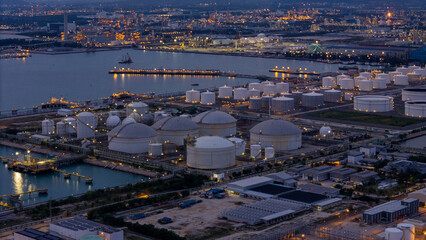 large industrial oil and gas storage tanks at a coastal refinery terminal during dusk. Tanker ships docked at long piers highlight the global energy transport infrastructure.
