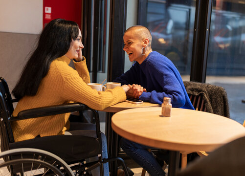 Two friends holding hands and enjoying a coffee break at a cafe, one of them using a wheelchair