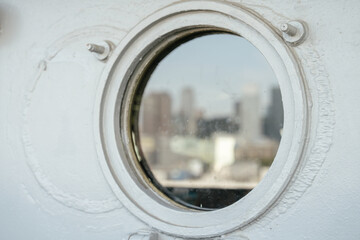 A circular porthole window on a white ship wall shows a soft, blurred view of a city skyline in the distance. The metal frame and bolts around the porthole are in sharp focus.
