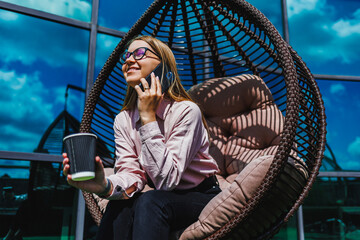 A woman relaxes in a modern hanging chair outdoors, surrounded by greenery. Sipping coffee and...