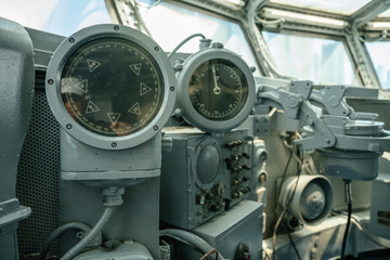 Close-up of vintage navigation instruments inside a ship's bridge. Compass dials and mechanical controls are surrounded by steel panels and wiring.