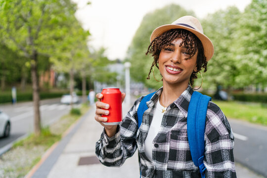Happy latin student holding a red soda can, smiling brightly while walking along a sunny city street, enjoying a carefree moment outdoors