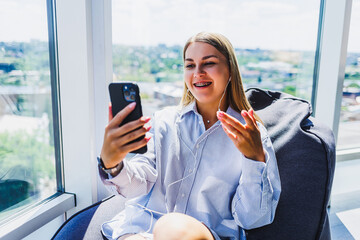 A young woman is happily engaged in a video chat while relaxing comfortably in a stylish, modern living space that features a beautiful and scenic view outside her window, creating a perfect ambiance