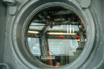 A circular porthole window reveals the interior of a ship's cockpit with visible wiring and equipment. The image captures reflections and light from both inside and outside, creating a layered visual