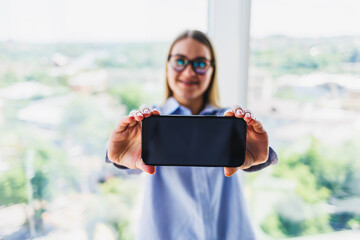A young woman, full of energy and enthusiasm, is holding a smartphone in a contemporary office setting that boasts a stunning scenic view, embodying the essence of modern business and connectivity