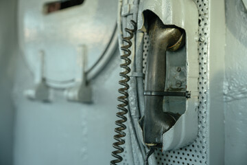 An aged black telephone handset is mounted on a metal wall aboard a military ship. The handset is...