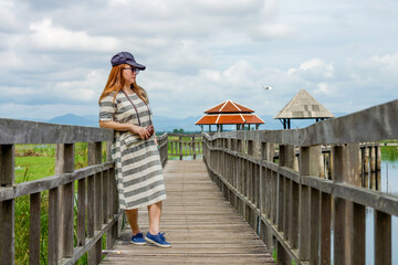 Woman Enjoying Scenic View at Bueng Bua Wood Boardwalk. Female tourist with camera stands on the rustic wooden boardwalk at Bueng Bua, Sam Roi Yot, taking in serene wetland landscape under cloudy sky