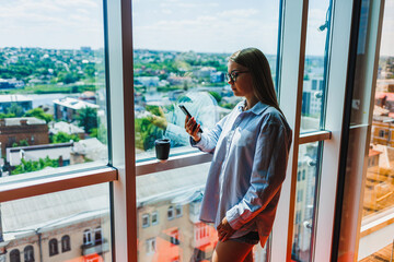 A young woman is reveling in the picturesque morning view from the window of a highrise apartment...