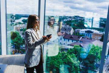 A confident woman in her modern office with a scenic view holds a smartphone, symbolizing her...