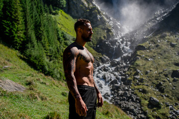 Naklejka premium Man tourist on summer vacation. Handsome man with naked torso on summer vacation day. Sexy hiker outside. Tourist man walking on the waterfall landscape. Man Traveling and nature.