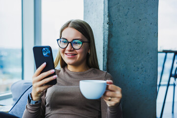 In a contemporary, stylish indoor environment, a young woman is fully immersed in enjoying her coffee while engaging with her smartphone, showcasing a perfect blend of leisure and modern lifestyle