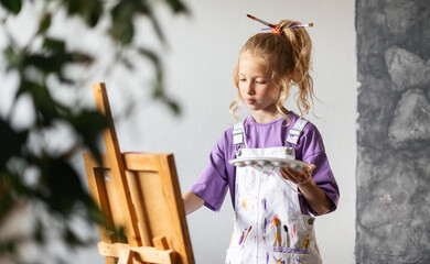 Little girl in paint-stained apron stands by easel holding palette and brush, learning to paint at home. Creative hobby helps kids develop artistic skills and imagination indoors.