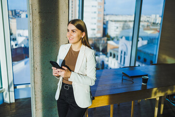A confident businesswoman is engaged with her smartphone in a modern office setting, showcasing her proficient skills in entrepreneurship and networking through digital means and technology