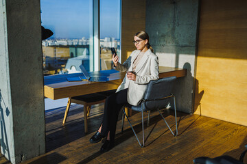 A chic and stylish businesswoman is enjoying her coffee while engaging in a phone interaction within a contemporary modern office setting that reflects elegance and professionalism