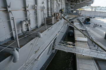The image shows the detailed side of a docked aircraft carrier with exposed pipes, mooring ropes, and a boarding gangway. The waterfront area includes safety barriers and modern pavement.