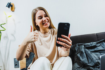 In a contemporary living room, a young woman, full of joy, sits comfortably on a stylish couch while holding her smartphone. She gives a thumbs up, clearly enjoying her modern, techsavvy lifestyle