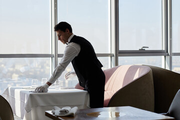 Caucasian young adult man arranging glassware on restaurant table wearing gloves, working as waiter...