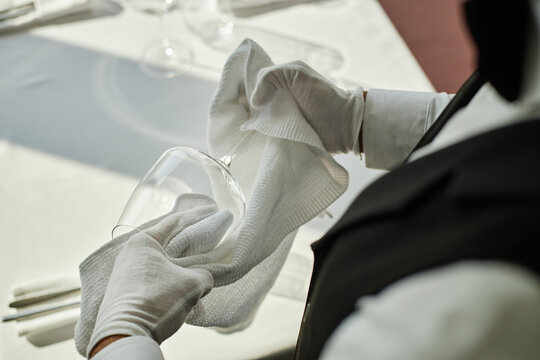 Unrecognizable waiter polishing wine glass with cloth wearing formal uniform and gloves in restaurant setting preparing table for service during work shift