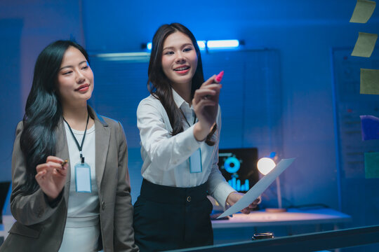 Businesswomen planning strategy, writing on glass board in office at night
