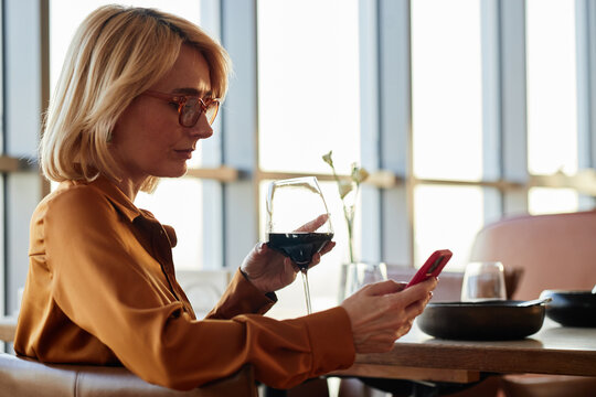 Middle aged Caucasian woman sitting at restaurant table holding wine glass in one hand and using smartphone with other hand, looking at screen with focused expression