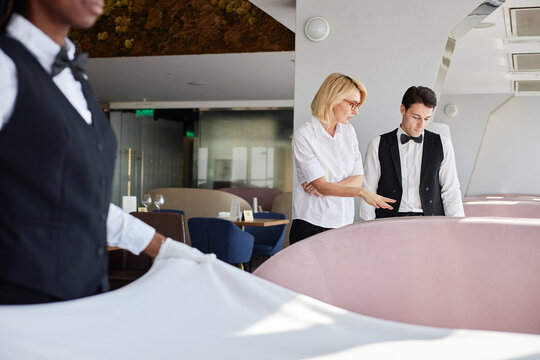 Caucasian middle aged woman instructing young adult Caucasian man in formal uniform while Black woman setting table in modern restaurant