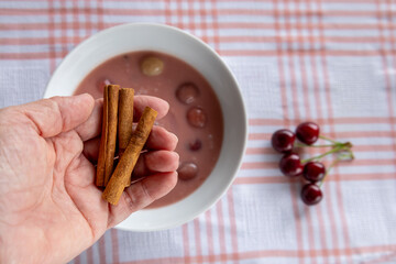 hand holding cinnamon sticks above cherry soup with fresh cherries on checkered tablecloth