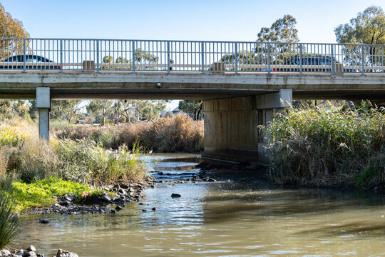 A road bridge spans the quiet Skeleton Creek with native grasses and reeds in Melbourne&rsquo;s outer suburbs. Cars cross above as shallow water flows beneath.Urban infrastructure and waterways in Australia