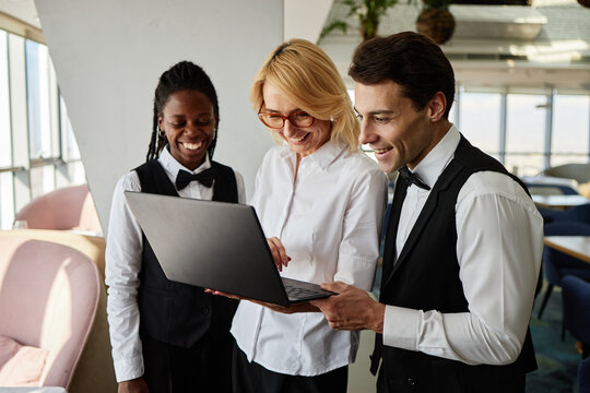 Caucasian middle aged woman standing with Black young adult woman and Caucasian young adult man using laptop together in restaurant, all smiling and wearing work uniforms