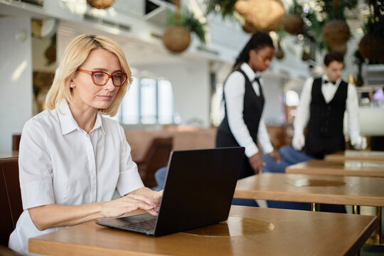 Caucasian middle aged woman working on laptop at restaurant table, Black woman and Caucasian man in uniforms preparing tables in background, focused on daily restaurant operations - Powered by Adobe