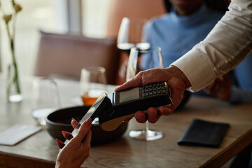 Male restaurant worker holding payment terminal while woman making contactless payment with smartphone at table, wine glasses visible