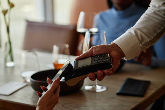 Male restaurant worker holding payment terminal while woman making contactless payment with smartphone at table, wine glasses visible
